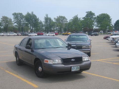 2001 Ford Crown Victoria - A dark gray police car is parked in a lot, with another police car behind it. The front of the car is visible.