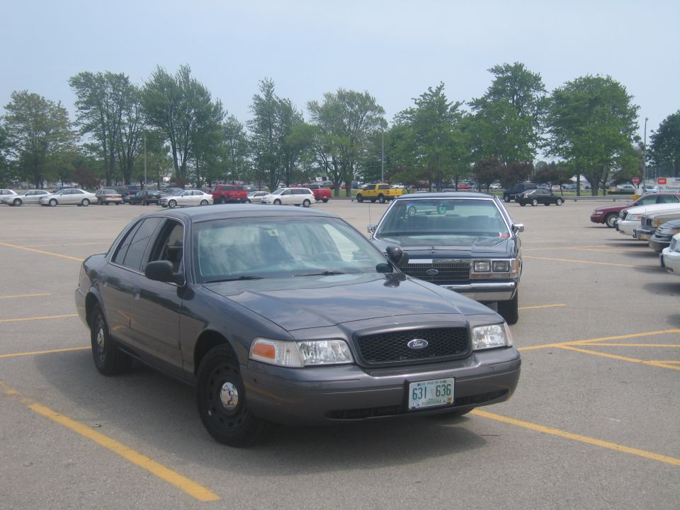 Dark gray police car parked in lot, another older police car behind it. License plate 631 636