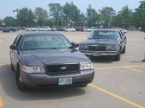 2001 Ford Crown Victoria - Two Ford Crown Victorias are parked in a lot, one dark gray and the other dark blue.