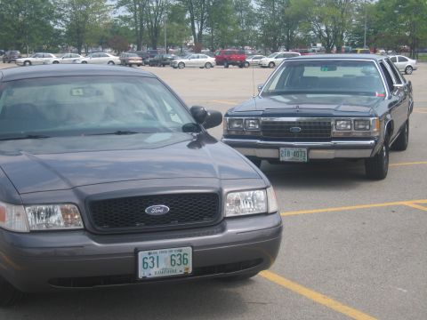 2001 Ford Crown Victoria - Two Ford Crown Victorias are parked side-by-side in a parking lot. The front vehicle is dark grey, and the rear vehicle is dark blue.