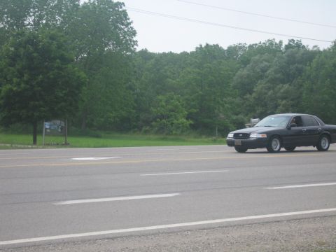 2001 Ford Crown Victoria - A black sedan drives down a two-lane road with a wooded area on the side.