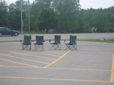 2001 Ford Crown Victoria - A blue sedan passes by a row of four folding chairs set up in a parking lot.