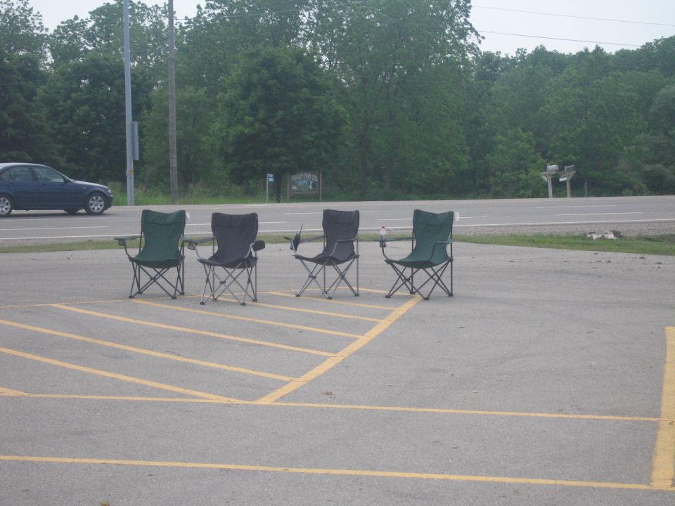 Four folding chairs in a parking lot with a blue sedan driving by on the road
