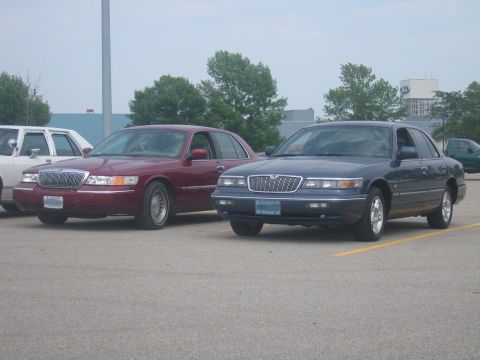 2001 Ford Crown Victoria - Two sedans are parked side-by-side in an outdoor parking lot under a cloudy sky.