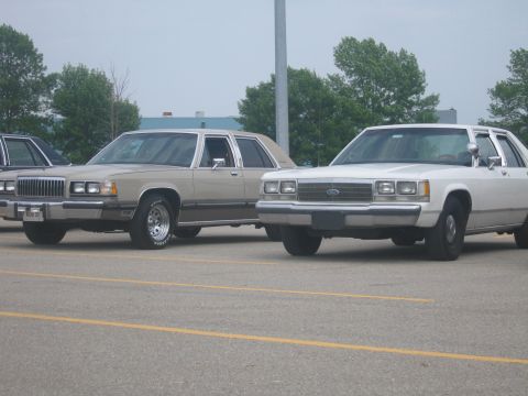 2001 Ford Crown Victoria - A tan sedan and a white sedan are parked in a lot, with the white car showing a Ford emblem.