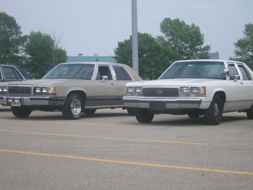 Two sedans, one tan and one white, parked side-by-side. The tan car has a prominent grille and chrome wheels. The white car has a Ford emblem on its grille