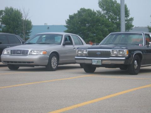 2001 Ford Crown Victoria - Two Ford Crown Victorias, a silver model and a black model, are parked side-by-side in a parking lot.