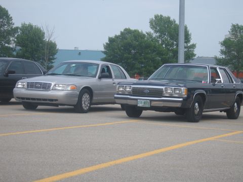 2001 Ford Crown Victoria - Two Ford Crown Victorias are parked in a lot, one dark blue and one silver.