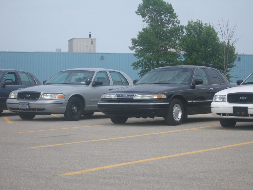 Three police sedans parked in a lot. The middle one is black, the one to its left is silver, and the one to its right is white
