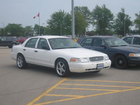 2001 Ford Crown Victoria - A white 2001 Ford Crown Victoria is parked in a lot next to other vehicles.
