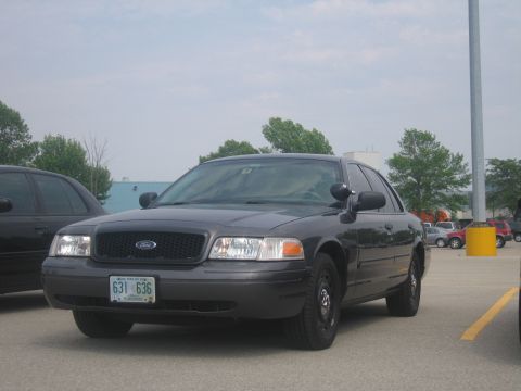 2001 Ford Crown Victoria - A dark gray Ford Crown Victoria police car is parked in a lot with a yellow pole and trees in the background.