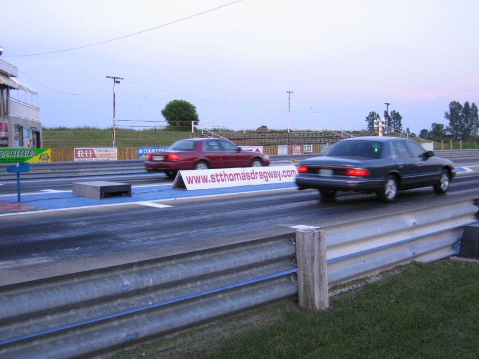 Two sedans racing down a drag strip past a stthomasdragway.com banner