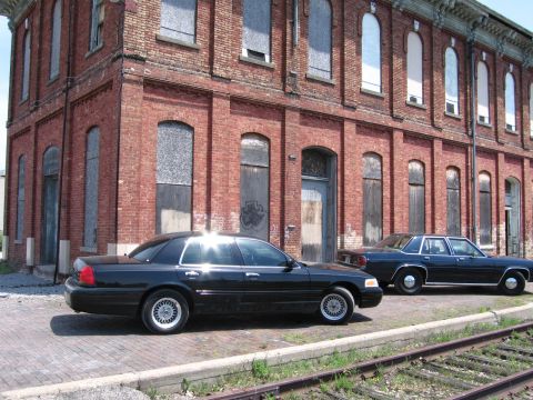 2001 Ford Crown Victoria - Photograph of Ford sedans beside a brick building and rail tracks, not a technical document.