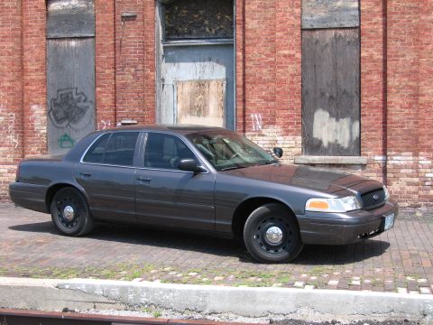 2001 Ford Crown Victoria - Photo of a gray Ford Crown Victoria sedan parked in front of a weathered brick building.
