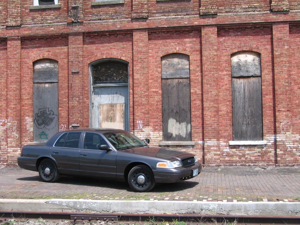 Gray Ford Crown Victoria sedan parked along railroad tracks at a brick wall