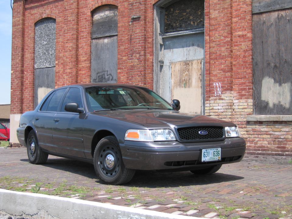 Gray Ford Crown Victoria sedan parked on brick pavement near a brick wall