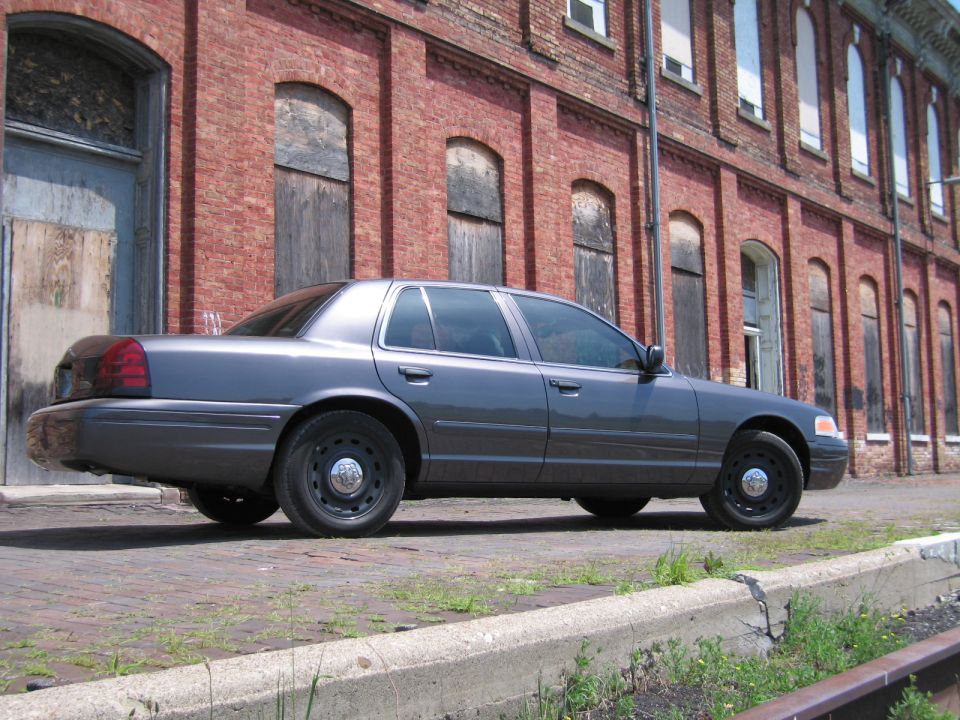 Rear three-quarter view of a gray Ford Crown Victoria sedan by a brick warehouse