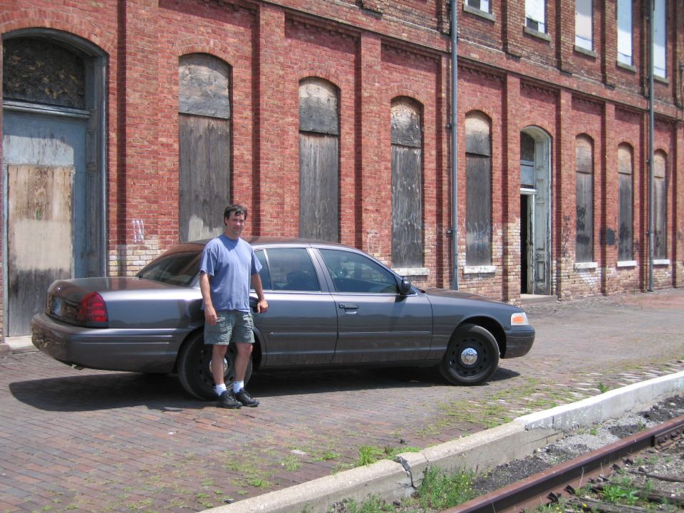 Man standing beside a gray Ford Crown Victoria sedan at a brick building