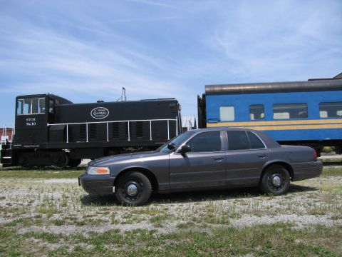 2001 Ford Crown Victoria - Photo of a gray Ford Crown Victoria sedan parked beside a black switcher locomotive and blue rail car.