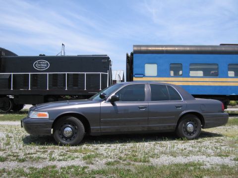 2001 Ford Crown Victoria - Photo of a gray Ford Crown Victoria sedan parked beside a black locomotive and blue passenger rail car.