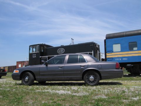 2001 Ford Crown Victoria - Photo of a gray Ford Crown Victoria sedan parked beside a black switcher locomotive and blue rail car.