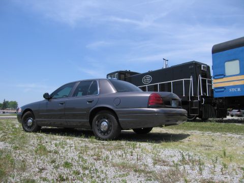 2001 Ford Crown Victoria - Photo of a gray Ford Crown Victoria sedan parked beside a black locomotive and blue rail car in a field.