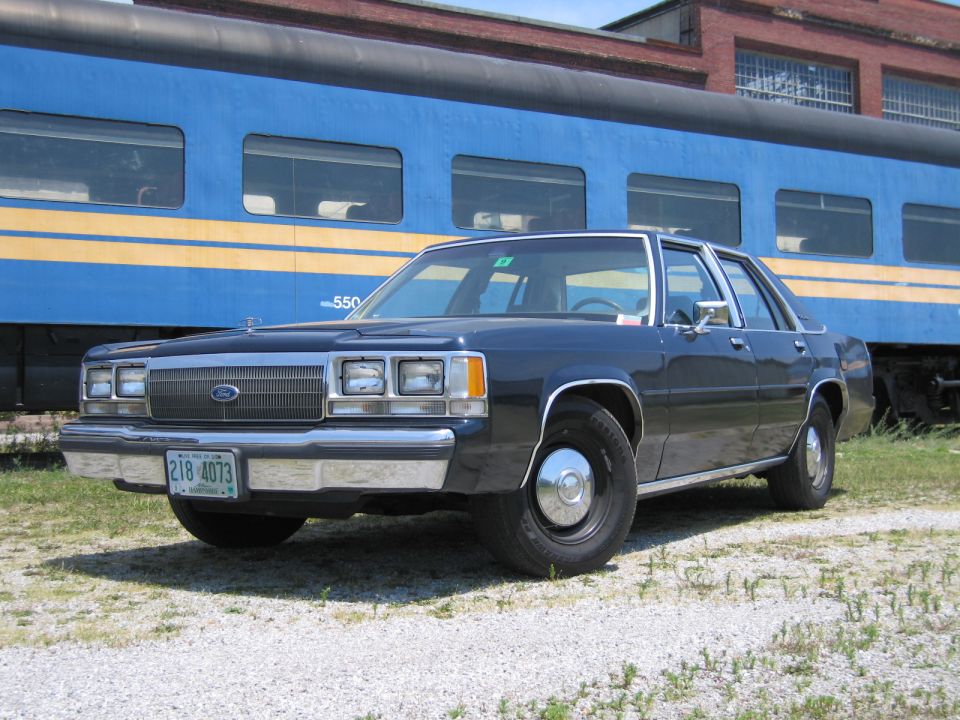 Black Ford LTD Crown Victoria sedan parked in front of a blue passenger rail car