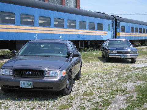 2001 Ford Crown Victoria - Photo of two Ford Crown Victoria sedans parked outdoors beside a blue passenger train car.