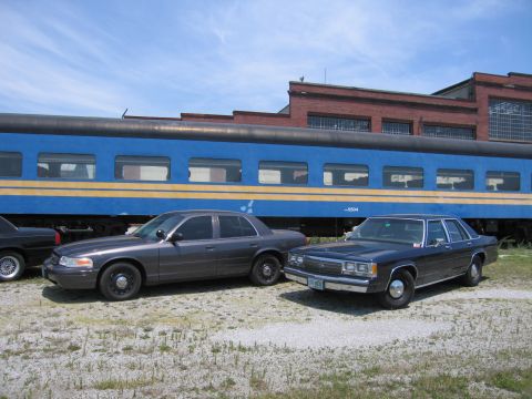 2001 Ford Crown Victoria - Photo of two Ford Crown Victoria sedans parked side by side in front of a blue passenger train car.