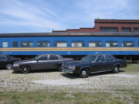 2001 Ford Crown Victoria - Photo of two Ford Crown Victoria sedans parked beside a blue STCR 5504 passenger train car.