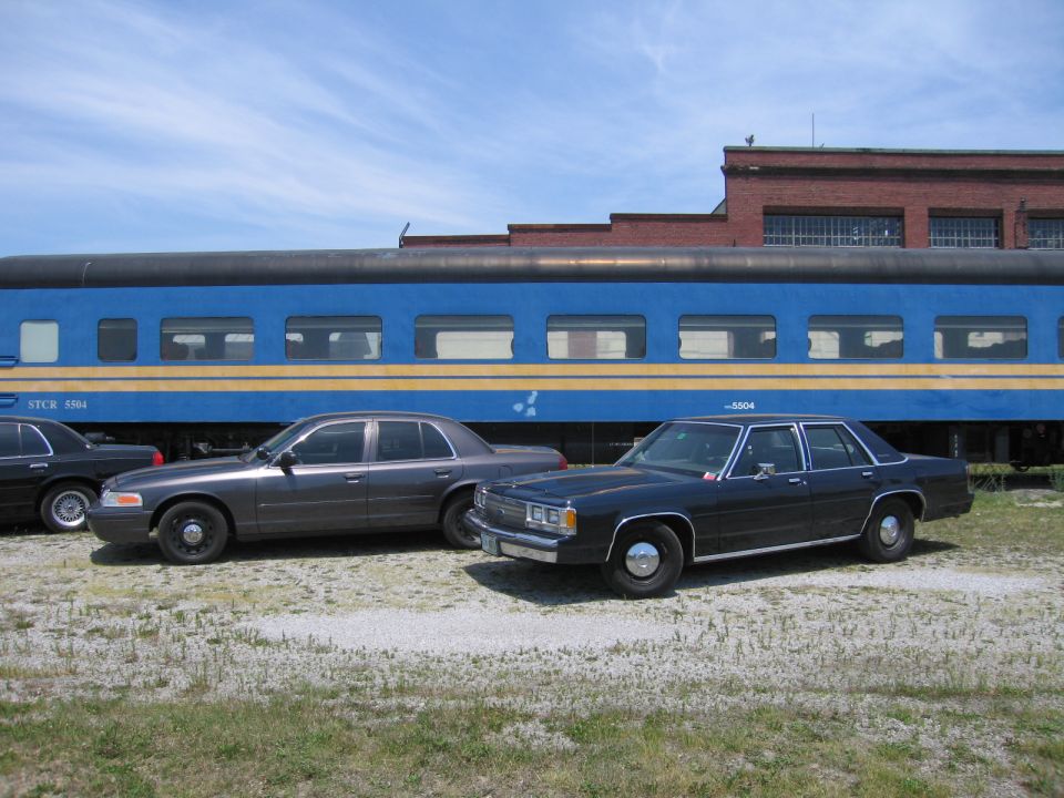 Two Ford sedans parked beside a blue STCR 5504 passenger rail car