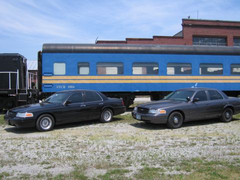 2001 Ford Crown Victoria - Photo of two black Ford Crown Victoria sedans parked beside a blue STCR 5504 passenger train car.