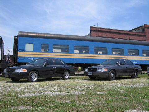 2001 Ford Crown Victoria - Photo of two Ford Crown Victoria sedans parked beside a blue passenger train car.