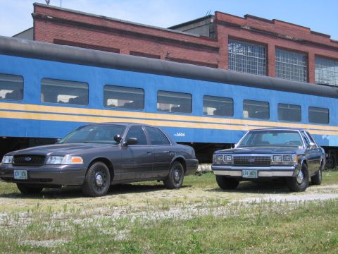 2001 Ford Crown Victoria - Photo of two Ford Crown Victoria sedans parked beside a blue passenger train car and brick building.