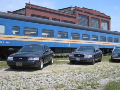 2001 Ford Crown Victoria - Photo of two Ford Crown Victoria sedans parked outdoors beside a blue STCR 5504 passenger train car.