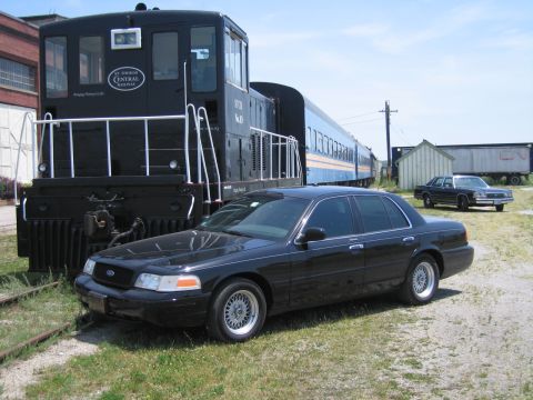 2001 Ford Crown Victoria - Photo of a black Ford Crown Victoria sedan parked beside a black railway locomotive and passenger cars.
