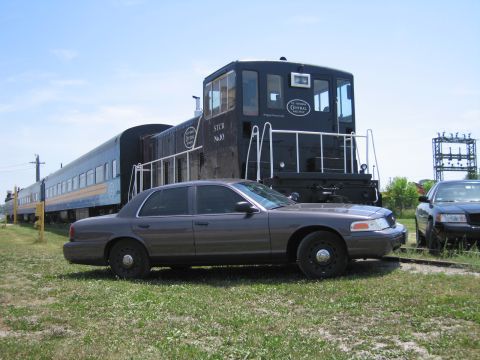 2001 Ford Crown Victoria - Side-profile photo of a gray Ford Crown Victoria sedan parked beside a black STCR railway switcher locomotive.