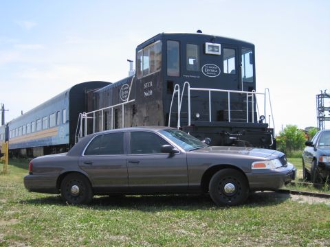 2001 Ford Crown Victoria - Photograph of a Ford Crown Victoria sedan parked next to a black diesel switcher locomotive on railroad tracks.