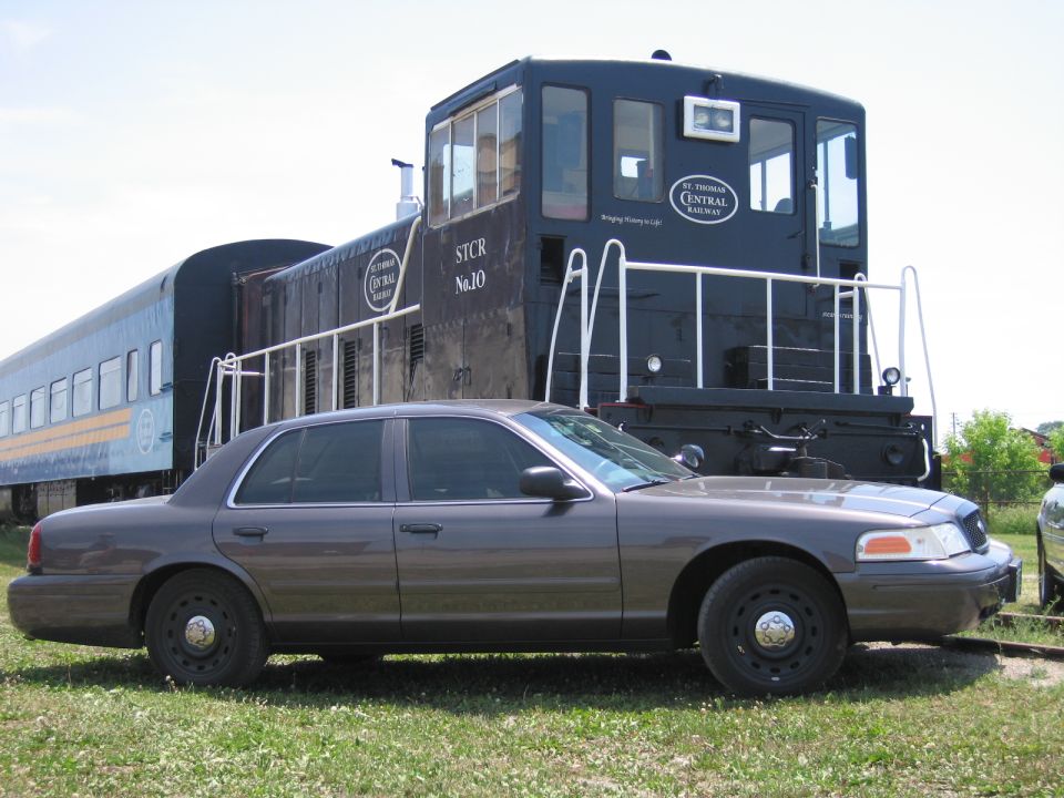 Side view of a gray Ford Crown Victoria in front of a black railway locomotive