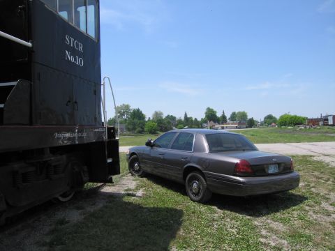 2001 Ford Crown Victoria - Photograph of a Ford Crown Victoria sedan beside the rear of a black railway locomotive in a railyard.