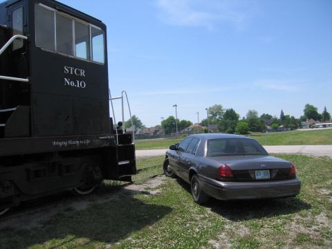 2001 Ford Crown Victoria - Photograph of a gray Ford Crown Victoria parked at the end of a black diesel railway locomotive.