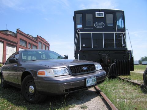 2001 Ford Crown Victoria - Photograph of a Ford Crown Victoria sedan parked next to a black locomotive near an old brick rail building.