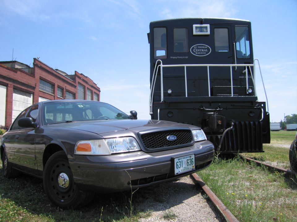 Front of a gray Ford Crown Victoria beside a black locomotive by a brick building