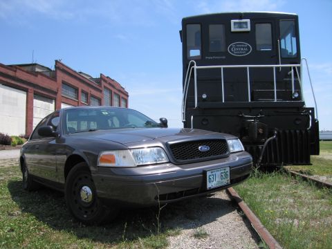 2001 Ford Crown Victoria - Photograph of a gray Ford Crown Victoria parked beside a black diesel locomotive on rail tracks.