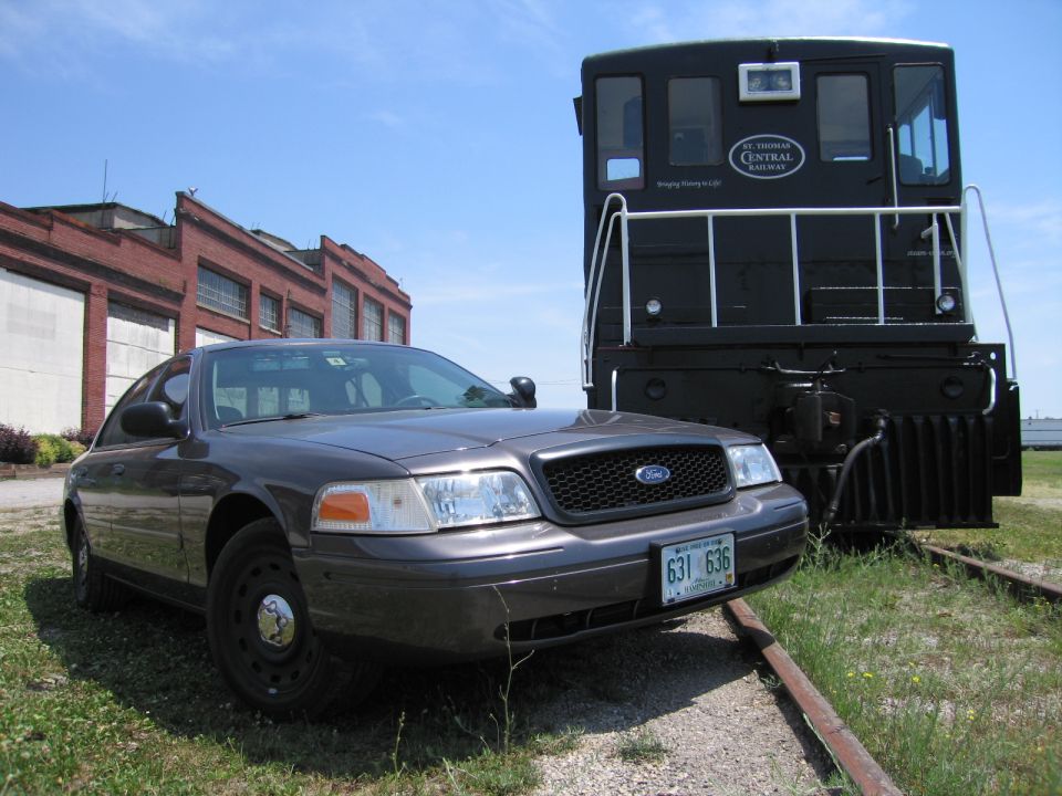 Gray Ford Crown Victoria parked front-on beside a black railway locomotive