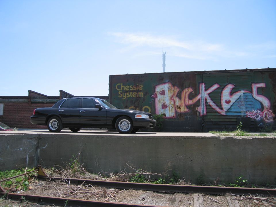 Black Ford Crown Victoria parked above railroad tracks near a graffiti boxcar