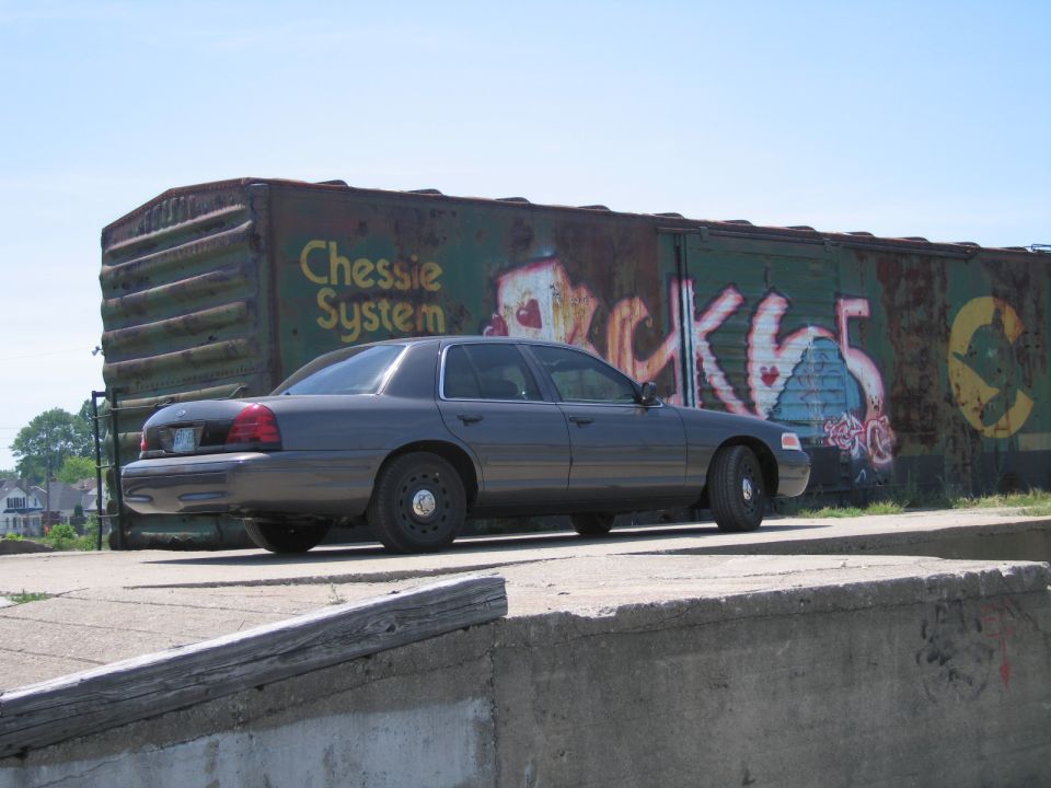 Gray Ford Crown Victoria parked atop a wall near a graffitied boxcar