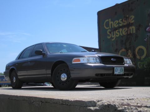 2001 Ford Crown Victoria - A dark sedan is parked outdoors under a clear blue sky, with a building to its right.
