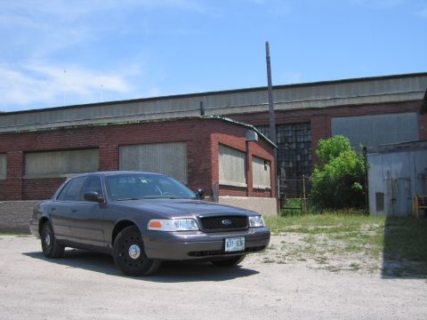 2001 Ford Crown Victoria - A gray Ford Crown Victoria police car is parked in front of an industrial brick building under a blue sky.