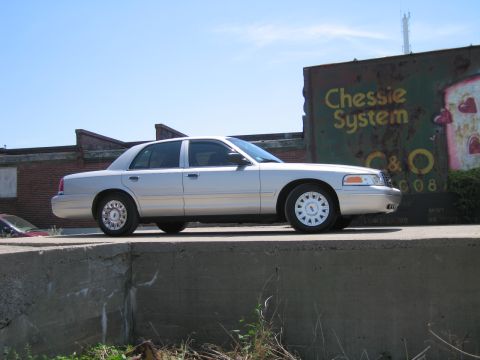 2001 Ford Crown Victoria - A silver sedan is parked on a concrete ledge with a weathered sign in the background.